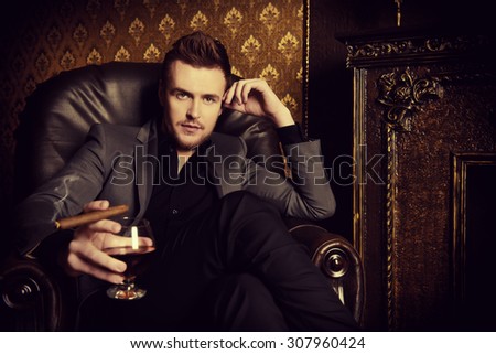Elegant man in a suit with glass of beverage and cigar in vintage room. Fashion.