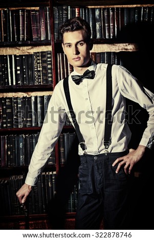Handsome well-dressed young man stands by bookshelves in a room with classic interior. Fashion shot.