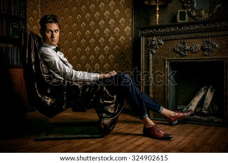 Elegant handsome young man sitting by the fireplace in a room with classic vintage interior. Fashion shot.