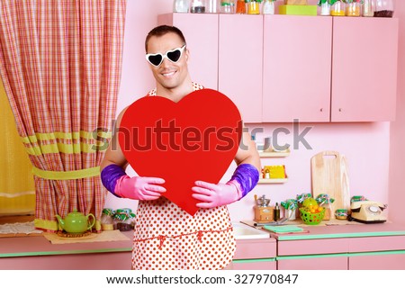 Handsome muscular man in an apron holding big red heart. Pink kitchen. Love concept. Valentine\'s day.