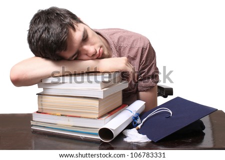 Male student sleeping on textbooks with graduation certificate and blue cap on a wooden desk