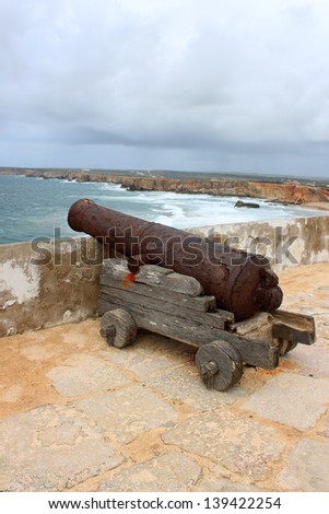 Rugged and rocky cliffs of the Algarve coastline in Sagres, Portugal, on the Atlantic Ocean under dark rainy sky and cannon showing protection