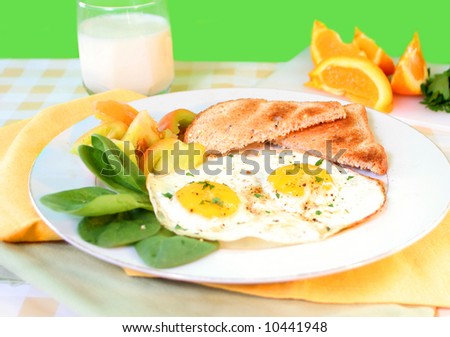 two sunny side up eggs on plate with fresh vegetables, whole wheat toast,  a glass of milk, and sliced orange in the background