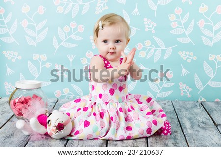 Girl in pink dress sitting on the wooden floor with candy jar on blue flower background holding a toy.