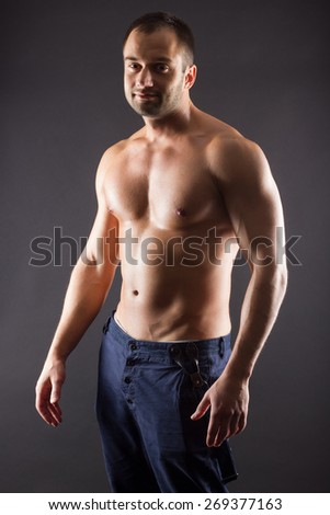 Portrait of sporty young muscular male topless shirtless athletic macho standing posing over black background. Torso of strong man in jeans against dark background.