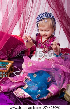 Little young east kid child  girl, sultana, Princess in Indian dress sari burqa scarf veil holding a pearl necklace sitting on a floor pillow at home interior under pink canopy. Childhood, copy space
