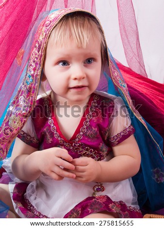 Little young east girl, sultana, Princess in Indian dress sari burqa scarf veil holding a pearl necklace sitting on a floor pillow at home interior under pink canopy. Childhood, copy space, doll house