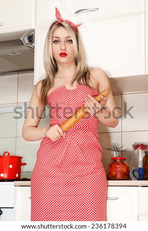 Portrait of young attractive caucasian blonde angry housewife with rolling pin at kitchen. Beautiful young housewife in the kitchen. Woman in pin up style.