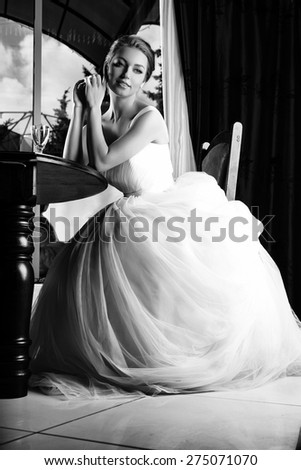 Beautiful blonde bride seats near table, is ready for a new bright life, inside interior. White Wedding dress. Pretty young woman love. Black and white photo.