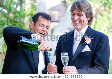 Gay couple pouring champagne to toast at their wedding.  The groom on the right is covered with champagne from the splash when the cork popped.