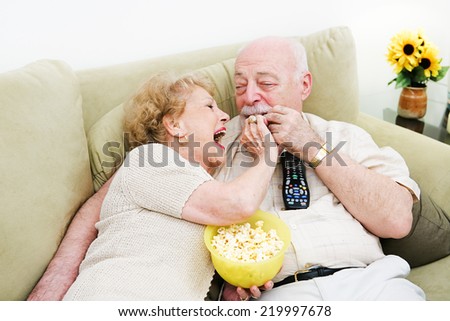 Senior woman laughs and feeds popcorn to her husband as they watch television.