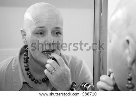 A woman bald due to a health issue, putting on makeup in the mirror.