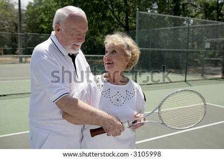 A senior couple playing tennis together.  He\'s showing her how to grip her racquet.