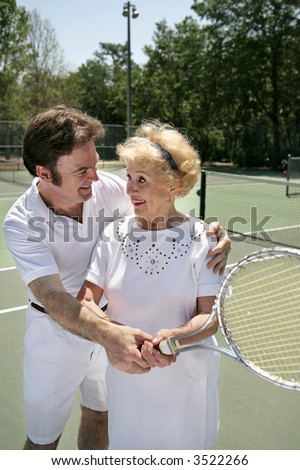 A pretty senior woman getting a tennis lesson from a handsome young pro.