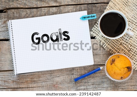 Notebook with goals sign and sticker remember on wooden desk with cup of coffee and muffin