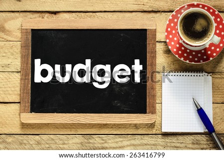 Budget on blackboard with coffee. Budget On blackboard with cup of coffee, notebook and pen on wooden background