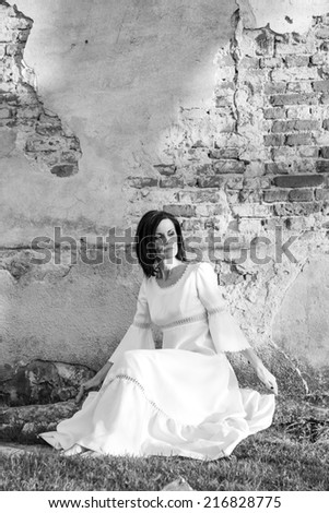 Beautiful, brunette woman in an old, abandoned house, wearing a wedding dress, posing with sunset light. Photo has grain texture visible on its maximum size. Artistic black and white photography