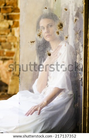 Beautiful girl shot through a dirty glass in an abandoned house wearing an old fashioned wedding dress. Photo has grain texture visible on its maximum size. Artistic photography