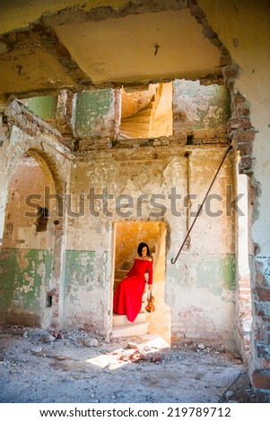 Beautiful, brunette woman in an old, abandoned house, wearing a red dress, looking sad and melancholic. Photo has grain texture visible on its maximum size. Artistic photography