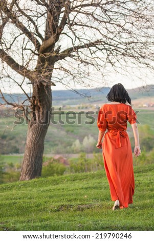Brunette girl posing in the field with tree, wearing an orange dress. Photo has grain texture visible on its maximum size. Artistic photography