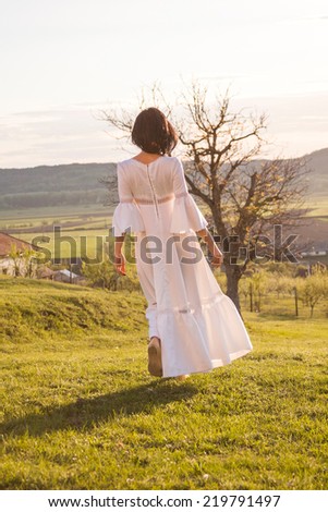 Brunette girl posing in the field with tree, wearing a white wedding dress posing with sunset light. Photo has grain texture visible on its maximum size. Artistic photography