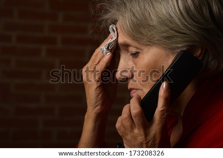 Older sick woman in red is speaking on phone on a background of a brick