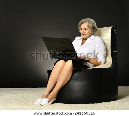 Smiling old woman sitting in business cloth on dark background with laptop