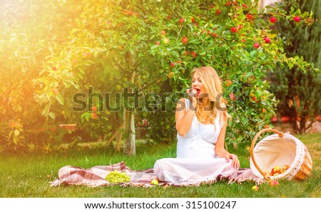 outdoor portrait of a pregnant woman in the garden on a plaid near apple tree, bite apple, with sun light vintage color