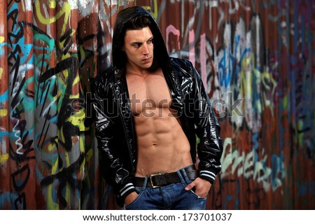 Young handsome macho man with open jacket revealing muscular chest and abs in industrial garage with graffiti .Fashion colors.