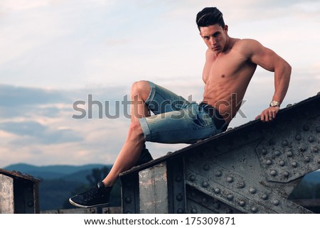 Handsome young muscle man shirtless with hand on rusty metal structure, looking in camera