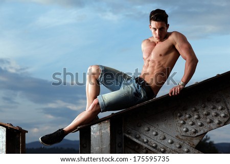 Handsome young muscle man shirtless with hand on rusty metal structure, looking in camera .Fashion photo