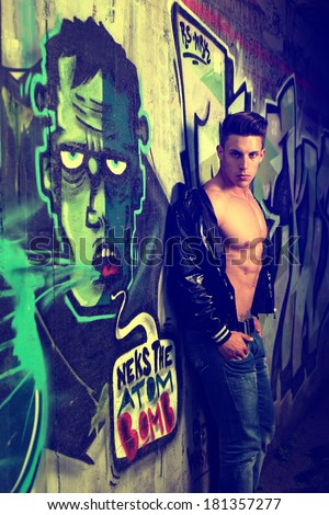 Young handsome macho man with open jacket revealing muscular chest and abs in industrial garage with graffiti .Fashion colors. No brands.