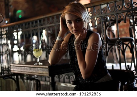 Attractive blonde woman  in elegant black dress sitting on bar stool. Gorgeous blonde model  posing provocatively in vintage bar