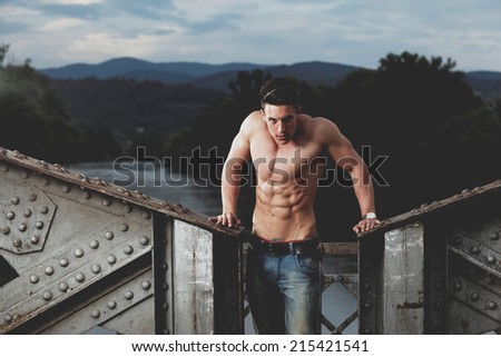Handsome young muscle man shirtless with hand on rusty metal structure, looking in camera .Photo has an intentional film grain) .Fine art.
