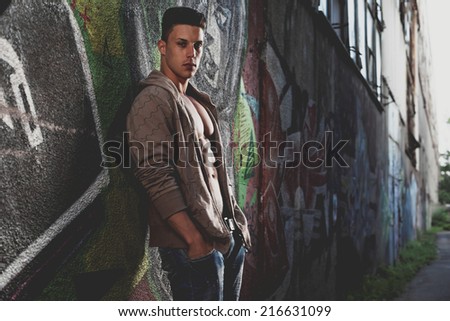 Young handsome macho man with open jacket revealing muscular chest and abs in industrial garage with graffiti