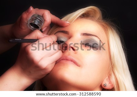 Make-up artist applying bright base color eyeshadow on model\'s eye and holding a shell with eyeshadow on background, close up