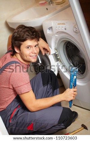 Young attractive smiling worker in uniform fixing washing machine, background