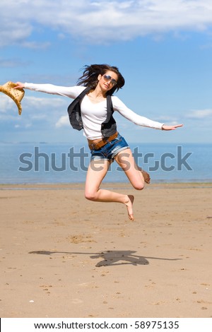 young attractive female jump high over sandy summer, wears sunglasses and hold straw hat in her hand