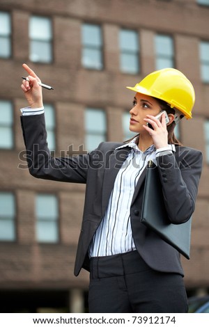 young female architect in hard hat talks over mobile phone and point to the building in front of her