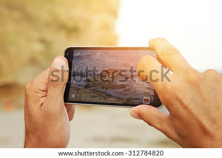 Close up on male hands taking photo of sea coast with mobile cell phone. Summer beach holiday vacation concept. Sunset colors
