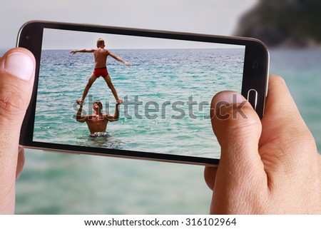 Male hand taking photo of man and his son playing in the sea with cell, mobile phone. Summer, holiday, vacation background