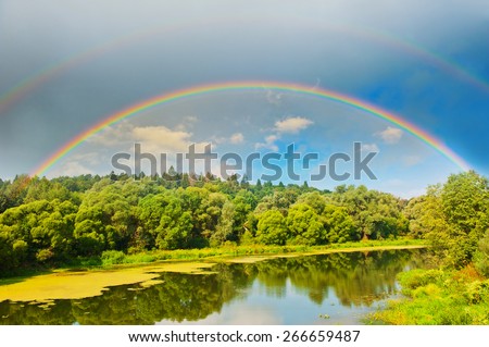 Bright rainbow in the sky with clouds above the forest and the river