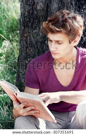Young man is sitting near the tree and reading a book