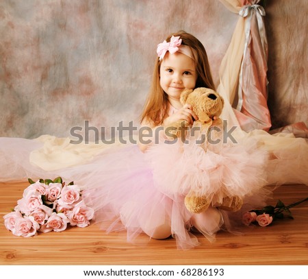 Adorable little girl dressed as a ballerina in a tutu, hugging a teddy bear sitting next to pink roses.