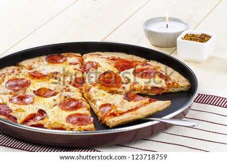 A pan of pepperoni pizza with serving spoon on a wooden table with lighted candle