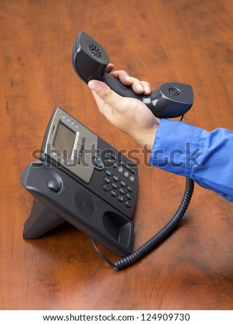 Close-up shot of human hand holding land line telephone receiver on wooden office desk.