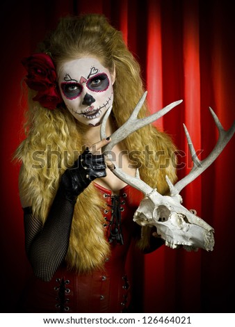 Portrait of a woman in traditional sugar skull holding a animal skull against red background.