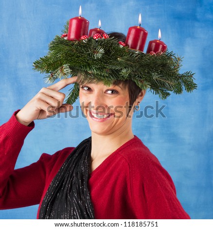 Smiling young woman woman wearing advent wreath on the head taps her forehead. Funny studio shot against a blue background, series