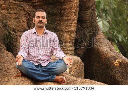 Close-up photo of a handsome indian executive doing meditation under a tree in a park. The person is sitting in lotus posture and is calm, relaxed closing his eyes for better concentration