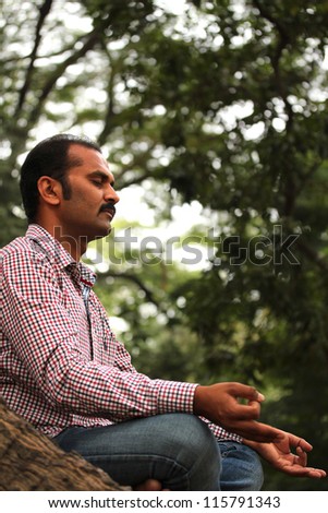 Close-up profile photo of a handsome indian businessman meditating under a tree in a garden. The person is sitting in lotus posture and is composed, relaxed closing his eyes for better concentration